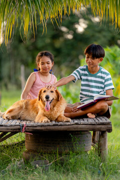 Vertical image of young Asian girl and boy as sister and brother relax on wooden litter also read some books with one dog sit beside and the look happy.
