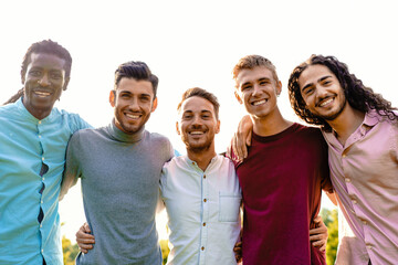 A multiracial group of five male friends embraces outdoors, sharing a moment of unity and joy. They...