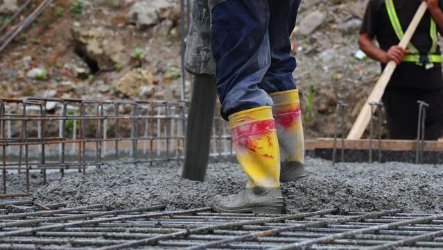 Worker pours concrete from a pipe onto the foundation of a building under construction, close-up of worker's feet.