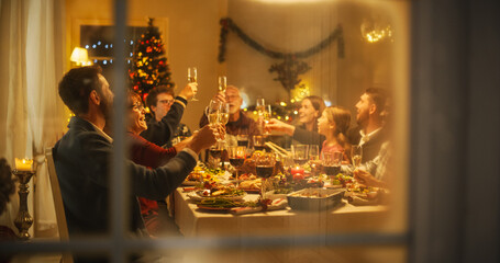 Cinematic Shot Through a Snowy House Window: Christmas Dinner Together with Parents, Children and Friends at Home. Multicultural Family Raising Champagne Glasses and Toasting, Celebrating Holidays