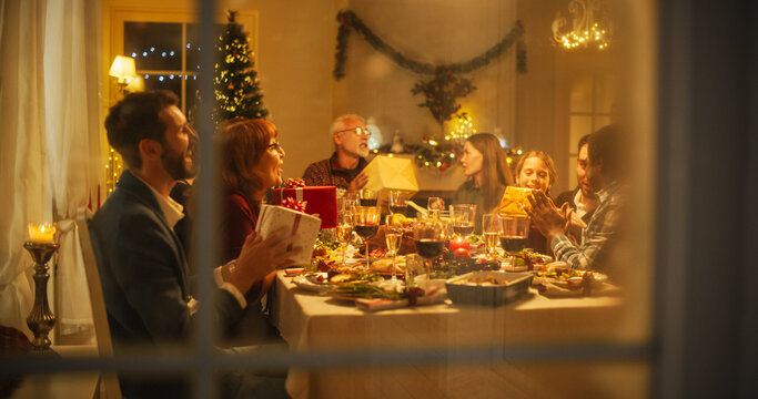 Snowy House Window: Diverse Family Exchanging Christmas Gifts To Celebrate Christmas. Beautiful Old And Young Relatives Excited To Receive Presents. Holiday Dinner Together At Home