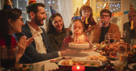 Family Celebrating Birthday with a Young Girl. Father is Holding a Celebratory Cake, Little Daughter is Blowing Out Candles and Making a Joyful Wish. Happy Child is Surrounded by Her Close Relatives