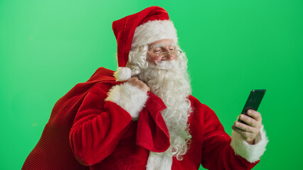 Studio Shot Isolated Mockup Template: Jolly Santa Claus Standing On Green Screen Chromakey Background And Holding Bag With Gifts, Using Mobile Smartphone. Christmas, New Year, Holiday Celebration