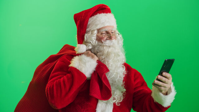 Studio Shot Isolated Mockup Template: Jolly Santa Claus Standing On Green Screen Chromakey Background And Holding Bag With Gifts, Using Mobile Smartphone. Christmas, New Year, Holiday Celebration - Powered by Adobe