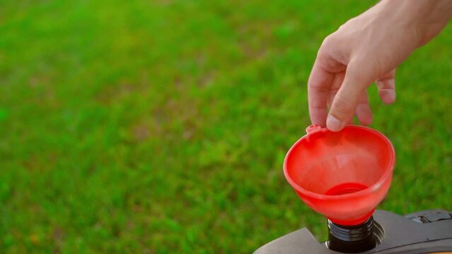 A full tank of fuel in the tank of a lawn mower against a background of a green lawn. A hand picks up a red plastic funnel after filling a lawnmower with gasoline, close-up