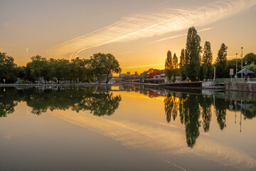 Obraz premium Paris, France - 08 15 2021: Ourcq Canal. Reflections on the Ourcq canal of a bridge, trees, barges and buildings at sunrise.