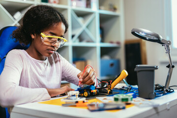 Smart Young African American Schoolgirl is Studying Electronics and Soldering Wires and Circuit Boards in Her Science Hobby Robotics Project. Girl is Working on a Robot in Her Room. Education Concept.