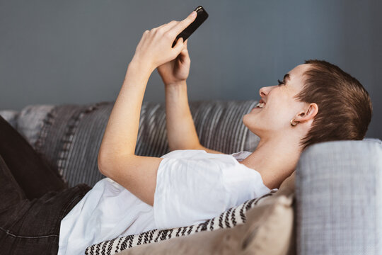 Young Modern Woman with Short Hair Relaxes on Sofa, Laughing at Smartphone