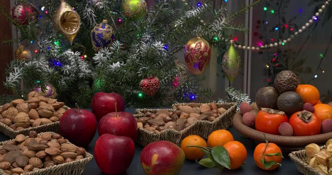 Assortment of healthy Christmas snacks: nuts and fresh fruits on a table with decorated tree. Flashing lights. Camera slide.