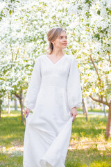 A young woman in a white dress walks against the backdrop of blooming apple trees. A girl posing against a background of flowers in a spring park.