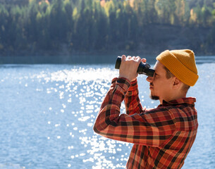 eco-conscious young man donning red plaid shirt, explores autumn forest by the side of tranquil...
