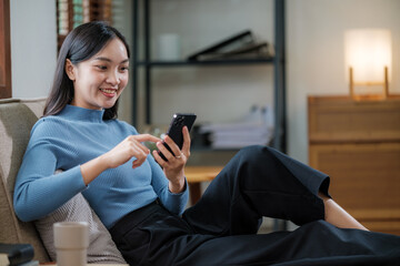 Happy relaxed young woman sitting on couch using cell phone, smiling lady laughing holding smartphone, looking at cellphone enjoying doing online e-commerce shopping in mobile apps or watching videos.