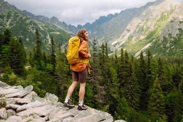 Naklejka premium Smiling traveler with a bright backpack on a hiking mountain trail. A beautiful woman on a cliff enjoying the mountain scenery. Active lifestyle.