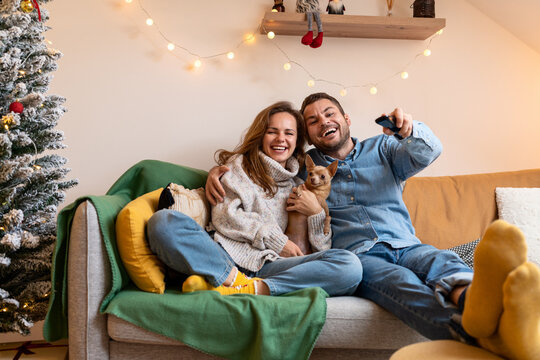 Happy young couple and their small dog sit on sofa at home and watching TV during Christmas holidays.