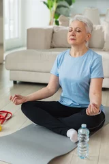 Schilderijen op glas Yoga mindfulness meditation. Senior adult mature woman practicing yoga at home. Mid age old lady sitting in lotus pose on yoga mat meditating relaxing. Older middle aged woman doing breathing practice © Юлия Завалишина