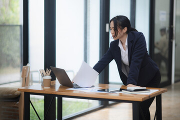 Asian woman working on laptop and standing at office desk.