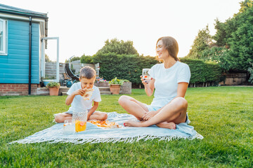 Happy Young mother and preschooler son having outdoor picnic dinner, eating pizza sitting on backyard Lawn on Sunny Day. Happy family time together. Active childhood. House in the suburbs in summer