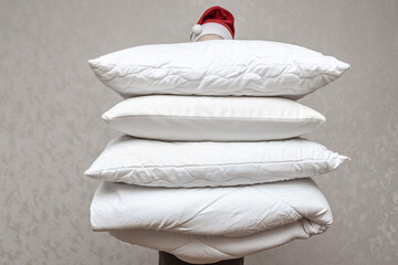Man in santa hat holding a warm duvet and feather pillows against a gray wall. Stack of bedding for...