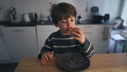Child eating piece of bread at kitchen table, adorable 4 year old boy snacking carb food on plate
