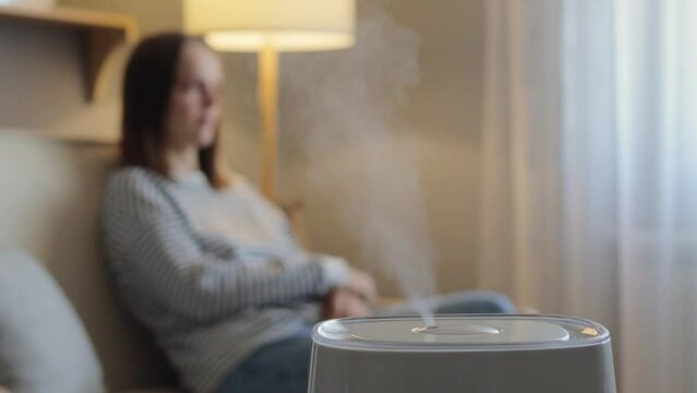 Air purifier in living room with young woman sitting relaxing on sofa caring for fresh air at home caring for well-being and health using air filter.