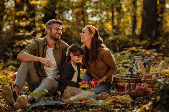 Mother, father and their daughter sitting on a blanket, holding apples and smiling in forest - Powered by Adobe