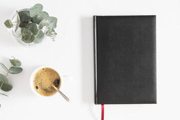 Top view of black closed cover blank notebook, pen, coffee cup, branch of green eucalyptus plant in vase on white table background. Creative workspace office. Business concept. Flat lay, top view