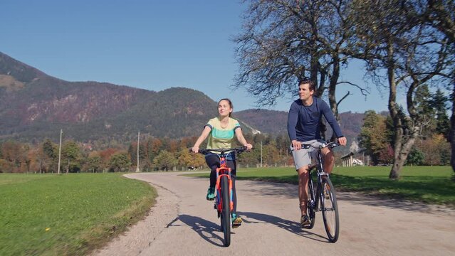 Young woman and a man riding bikes and enjoying beautiful nature, mountains, and colorful forests on a sunny fall day, front view. Country cycling and cycle tourism concepts.