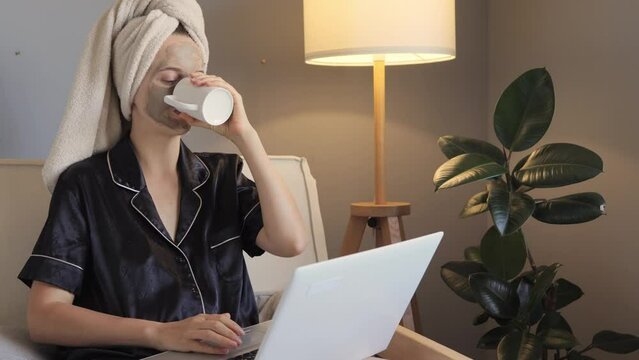Female freelancer at home taking care of her face and hair while working on laptop in living room and drinking coffee or tea browsing web pages wearing black pajamas and wrapped in towel.