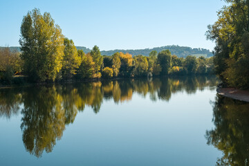 The tree lined banks of the Dordogne river in autumn colours reflected in the mirror calm water at...