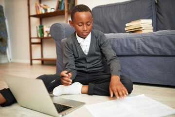 Side view indoor image of african american teen kid boy in school uniform study at home sitting on floor among papers and laptop, next to sofa, doing homework, studying or passing test online