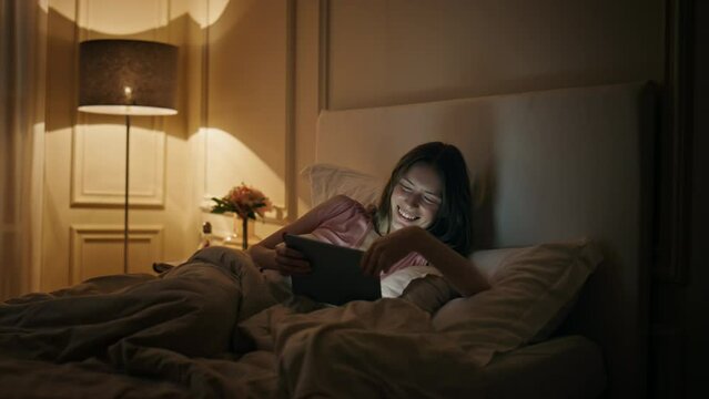 Cheerful woman watching tablet in bed. Happy female laughing movie in evening.