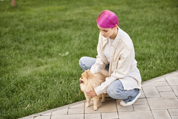 Fototapeta premium stylish woman with purple hair embracing cute pomeranian spitz on walkway in park near green lawn