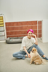 joyful pet hotel worker sitting on floor near pomeranian spitz and looking at camera in pet hotel