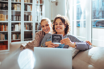 Happy young lesbian couple showing the x-ray of their future newborn on the laptop video call at home