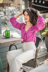 Happy Brunette Girl with pink blouse Sitting at the bar , Drinking a glass of lemonade While Smiling at the camera. Young sexy pretty woman on the chair  drinking  juice wearing a white pants