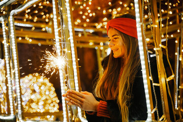 Woman holding sparkler night while celebrating Christmas outside. Dressed in a fur coat and a red headband. Blurred christmas decorations in the background. Selective focus