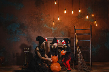 Mom and daughters in witch costumes sit near the fireplace and play with pumpkins.