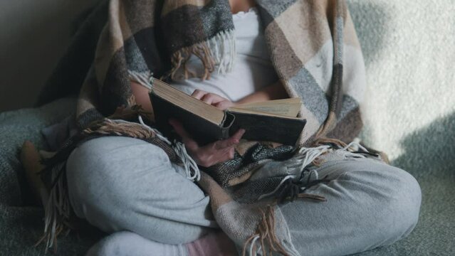 Female hands turning pages. Woman under cozy blanket reading book. Close up.