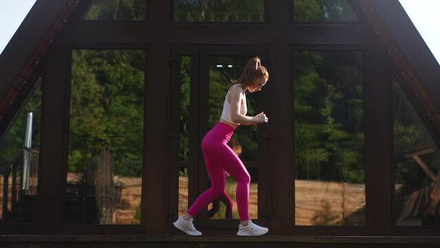 Silhouette of athletic female wearing sportwear rotating neck warming up before morning exercise, muscles warm up to prevent injury from workout, standing on background of bright sunlight, slowmo.