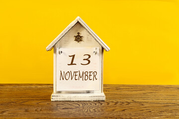 Calendar for November 13: the name November in English, numbers 13 on a decorative house on a wooden table, yellow background