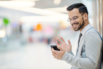 A happy young elegant man is using his phone for a video call while standing indoors and smiling at the phone.
