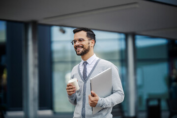 Fototapeta premium A happy young trendy businessman in smart causal is walking on the street with coffee to go and a laptop in his hands.