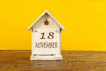 Calendar for November 18: the name November in English, numbers 18 on a decorative house on a wooden table, yellow background