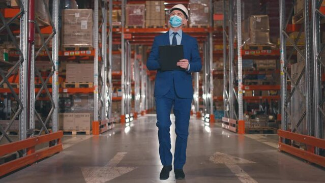 Manager walking along shipping stock. Head of company in official suit, protective helmet and safety mask. Inspector conducting audit at warehouse. Male hold folder at background of racks with boxes.
