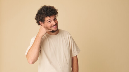 Smiling guy with curly hair dressed in beige t-shirt, shows call me phone hand sign gesture on beige background in the studio