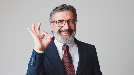 Middle-aged businessman in glasses with gray hair, shows ok gesture looking at camera, isolated on gray background in the studio