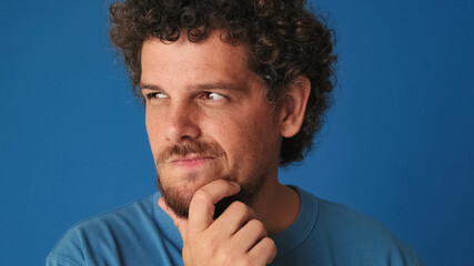 Fototapeta premium Close up, guy with curly hair dressed in blue t-shirt is thinking over difficult choice, imagining plan in his mind, having an idea, isolated on blue background in the studio