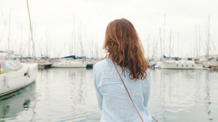 Attractive slim middle-aged woman sitting in the port looks into the distance at the yachts, back...