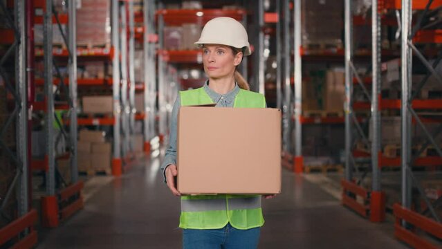 Female storage employee working at warehouse. Industrial worker carrying box at background of multi tiered shelves. Woman in protective helmet and reflective vest laying out goods. Logistic business.