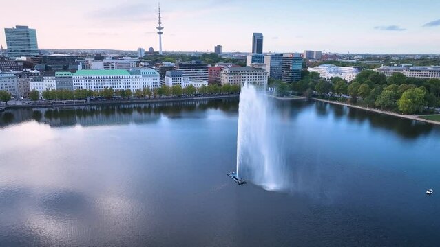 Aerial orbit of Hamburg's Alster Lake, where the Alster fountain takes center stage, surrounded by the impressive backdrop of the iconic Hamburg skyline and the bustling urban life. 